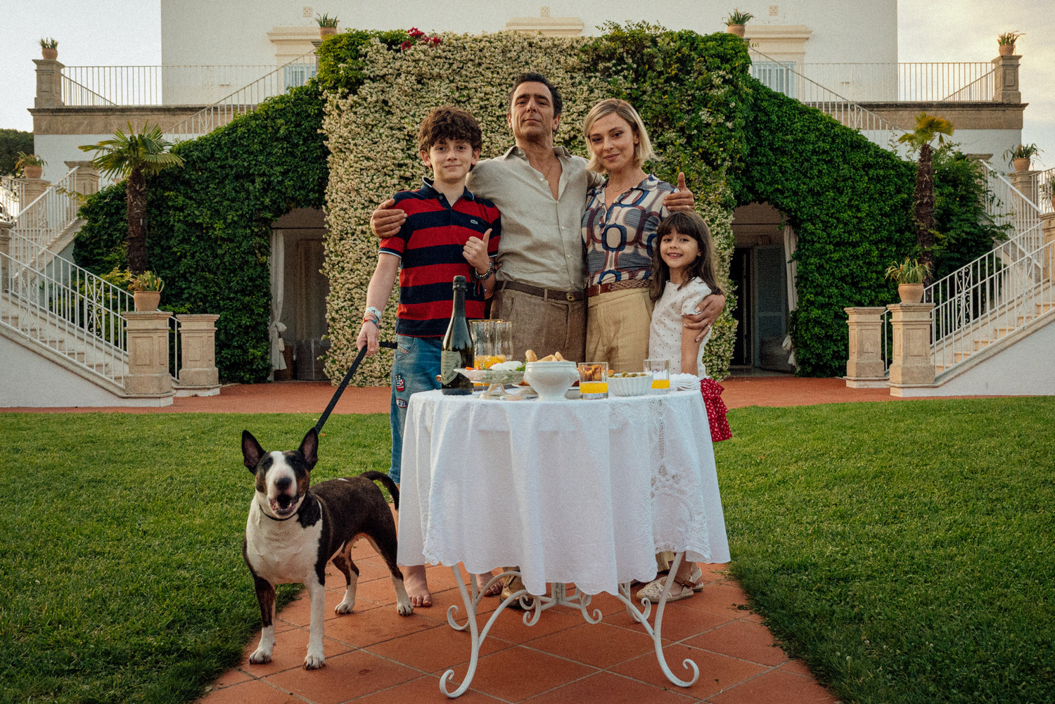 Scena de L’estranea di Paolo Strippoli: una famiglia in posa davanti a una villa ricoperta di edera, con un tavolo apparecchiato e un bull terrier.